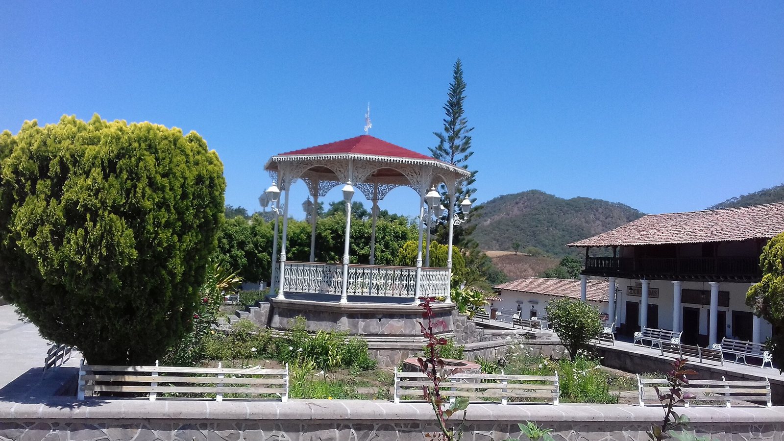 Kiosco blanco con techo rojo en la plaza de San Sebastián del Oeste, rodeado de jardines y bancas