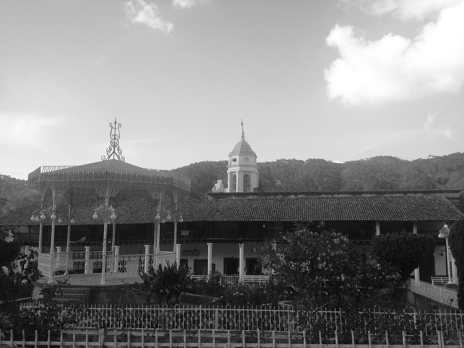 Fotografía en blanco y negro de la plaza de San Sebastián del Oeste con el kiosco, portales y la torre de la parroquia
