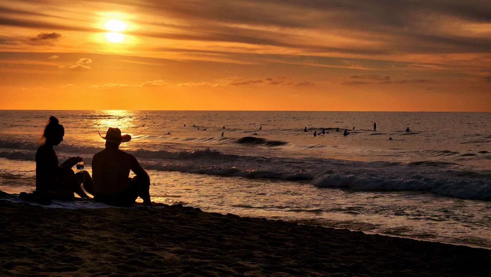 Siluetas de una pareja sentada en la arena al atardecer mientras varios surfistas esperan olas en San Pancho