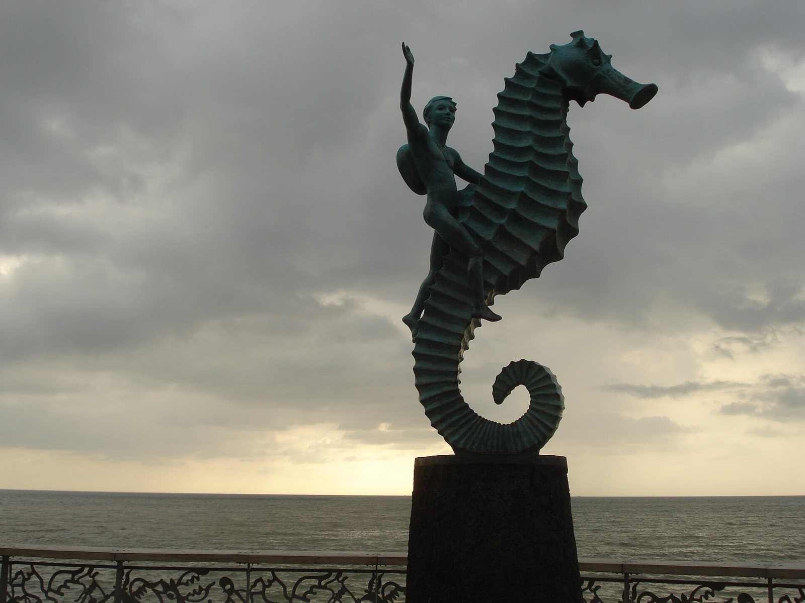 Escultura de bronce de un niño montado en un caballito de mar sobre un pedestal, con el mar y un cielo nublado detrás