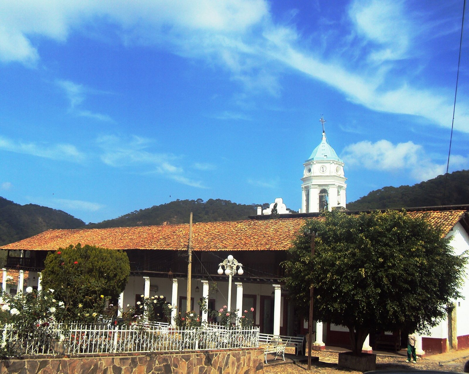 Torre blanca de la parroquia de San Sebastián sobre techos de teja, con la sierra cubierta de bosque al fondo