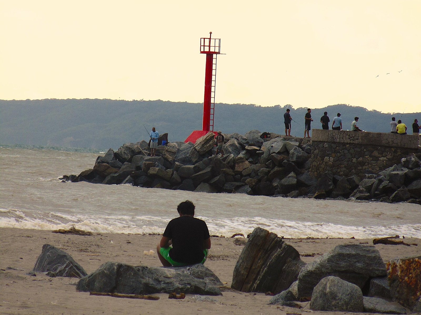 Faro rojo sobre la escollera de la marina, con personas pescando y la sierra al fondo