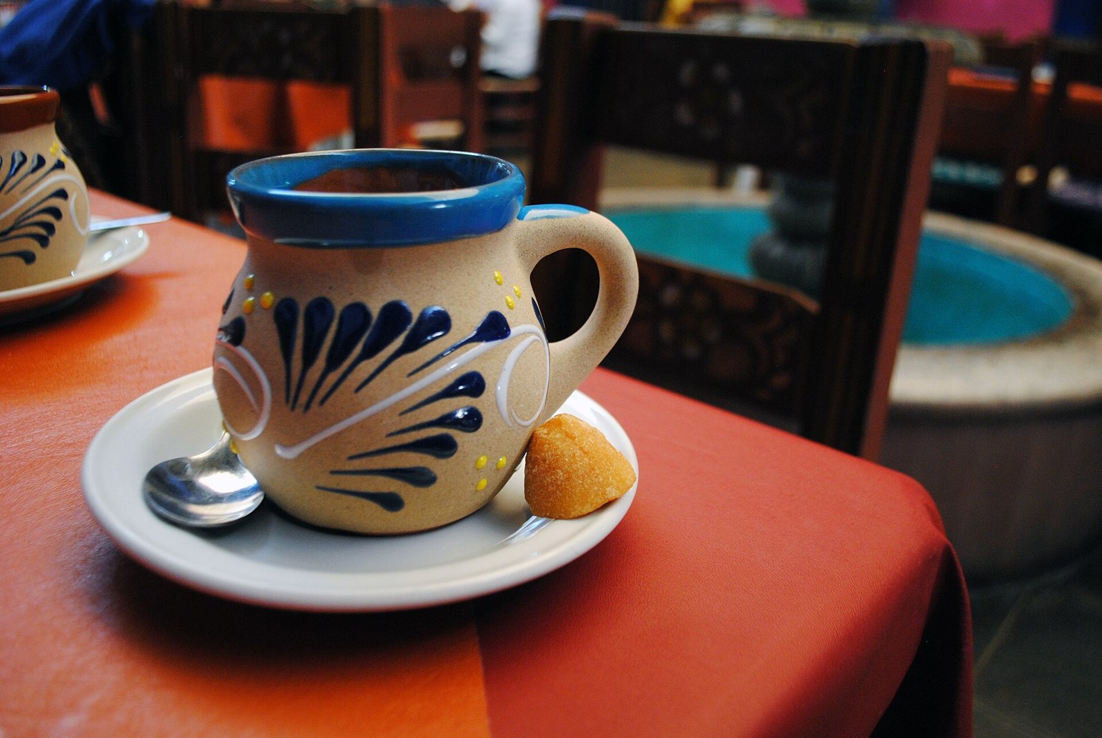 Jarrito de barro pintado con café, sobre plato con cuchara y un trozo de piloncillo