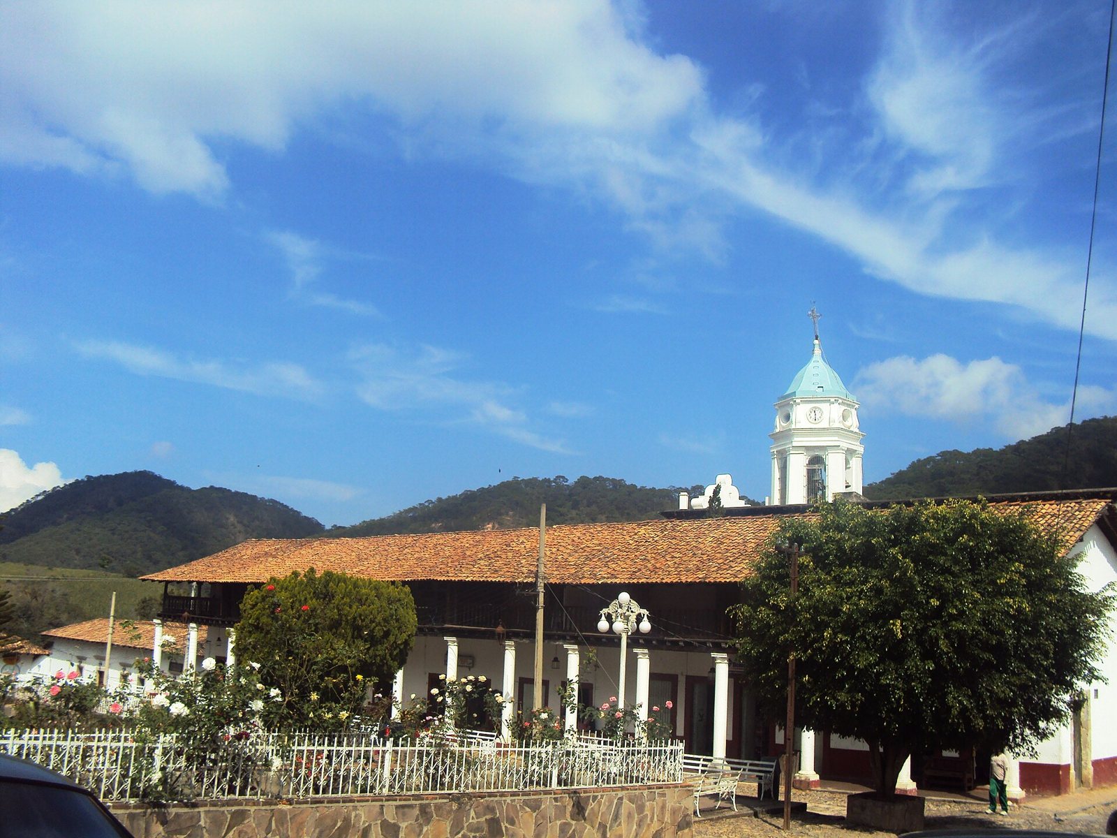 Torre blanca del templo de San Sebastián detrás de un edificio de portales con techo de teja, jardín con rosales y cerros con bosque al fondo