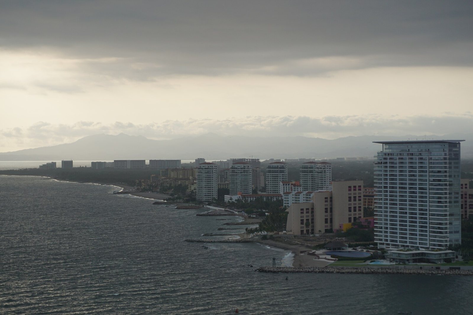Costa de Bahía de Banderas con edificios frente al mar y, detrás, la silueta de las montañas de la sierra bajo nubes grises