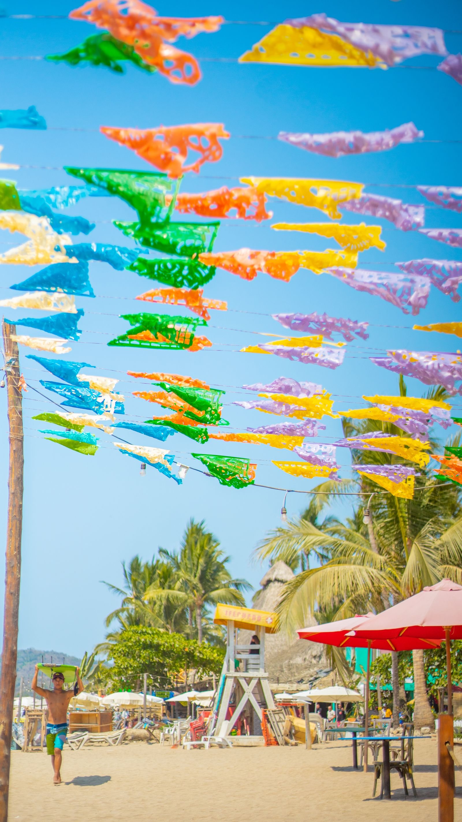 Papel picado de colores sobre la playa de Sayulita, con sombrillas, palmeras y una torre de salvavidas al fondo