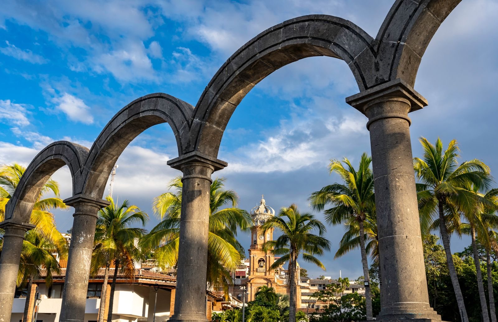 Arcos de piedra gris del Malecón de Puerto Vallarta enmarcando la torre con corona de la parroquia de Guadalupe, entre palmeras y techos de teja
