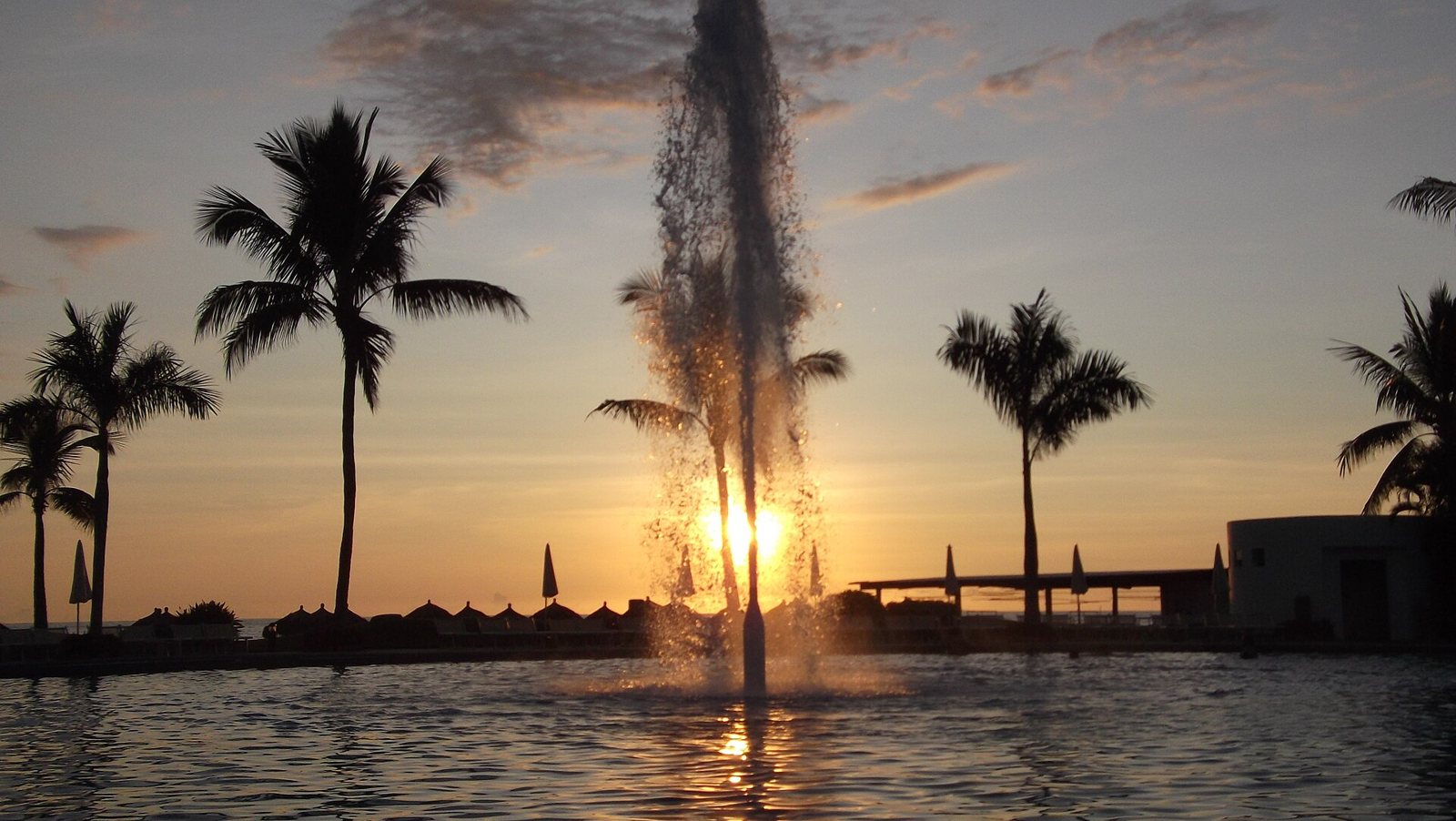 Fuente de agua en una alberca al atardecer, con palmeras en silueta y el sol sobre el mar en Nuevo Vallarta
