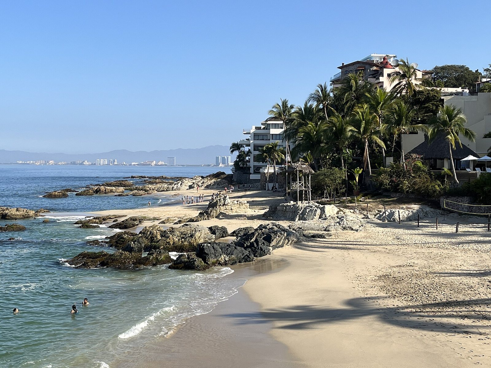 Playa de arena clara con rocas oscuras, palmeras y edificios sobre la ladera, con la costa de la bahía de Banderas al fondo