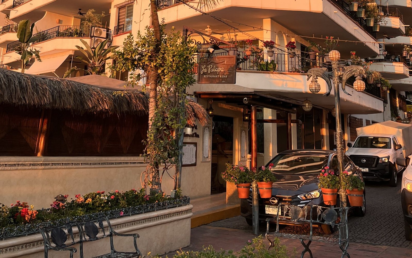 Entrada de La Palapa Restaurant & Bar con techo de palma, macetas con flores y luz dorada del atardecer en la calle Púlpito