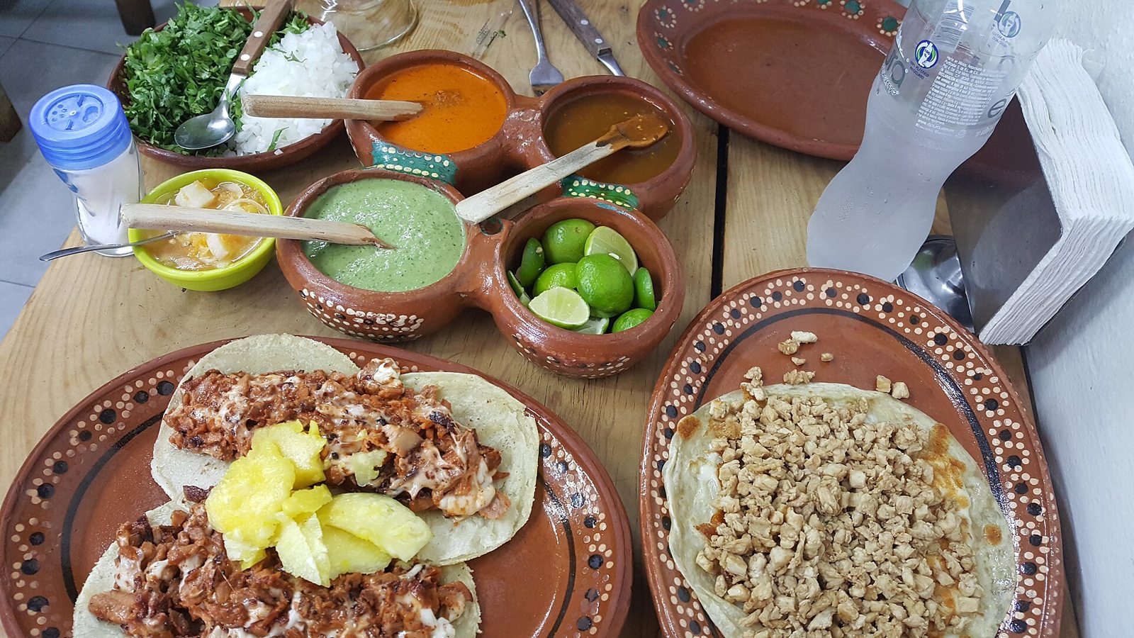 Tacos al pastor con piña en plato de barro, junto a salsas, limones, cilantro y cebolla en una mesa de madera