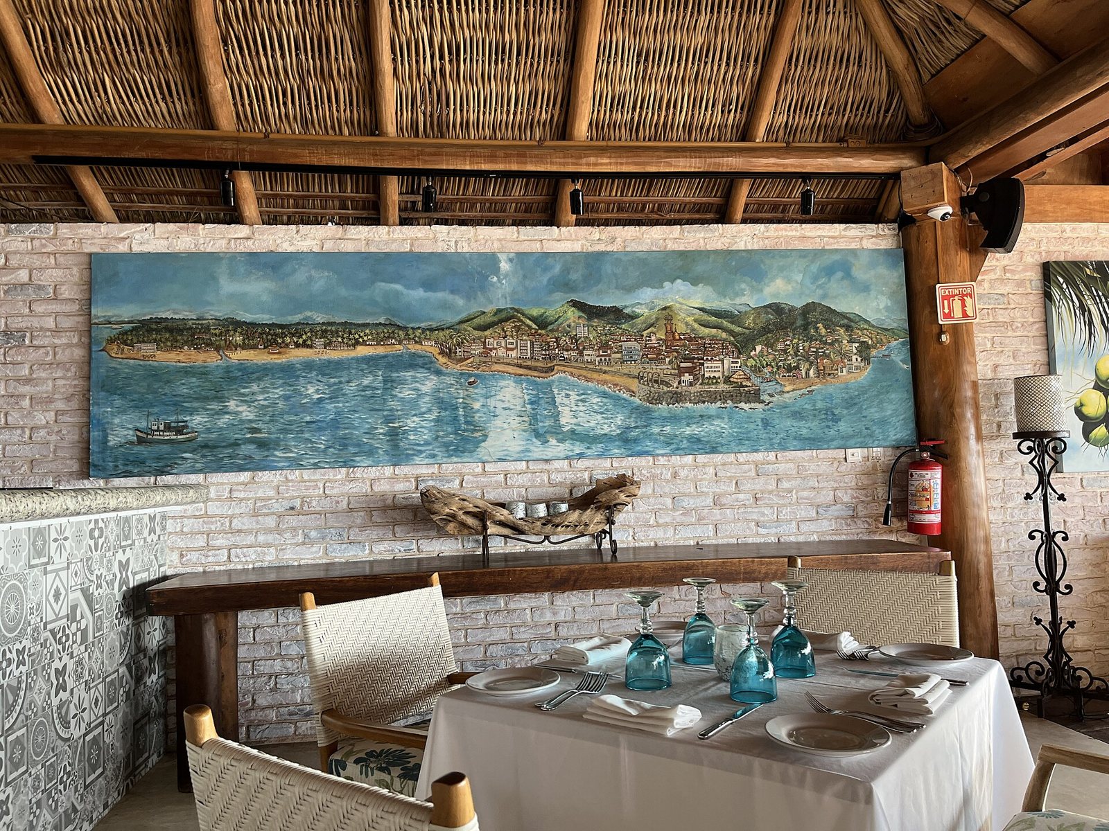 Mesa con mantel blanco y copas de vidrio soplado azul bajo un techo de palapa, con un mural que muestra la bahía y el pueblo de Puerto Vallarta