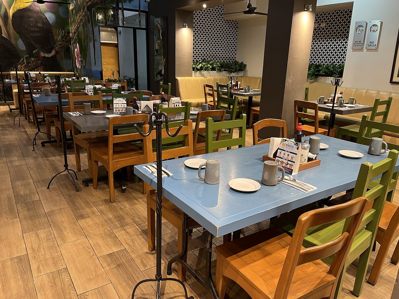 Comedor de Fredy's Tucán con mesas azules, sillas de madera verdes y naranjas, tazas de café y un mural de tucán en la pared