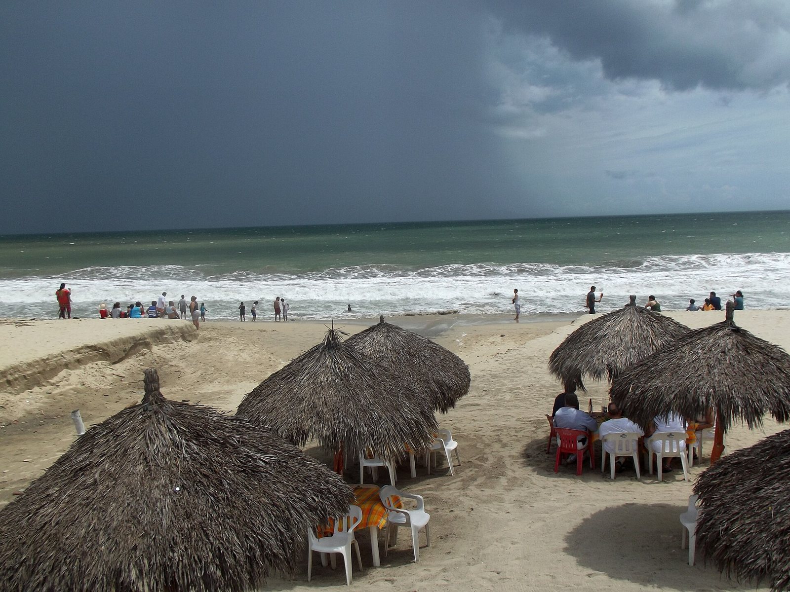 Sombrillas de palapa con mesas y sillas sobre la arena de la playa de Bucerías, con bañistas en la orilla y nubes de tormenta sobre el mar