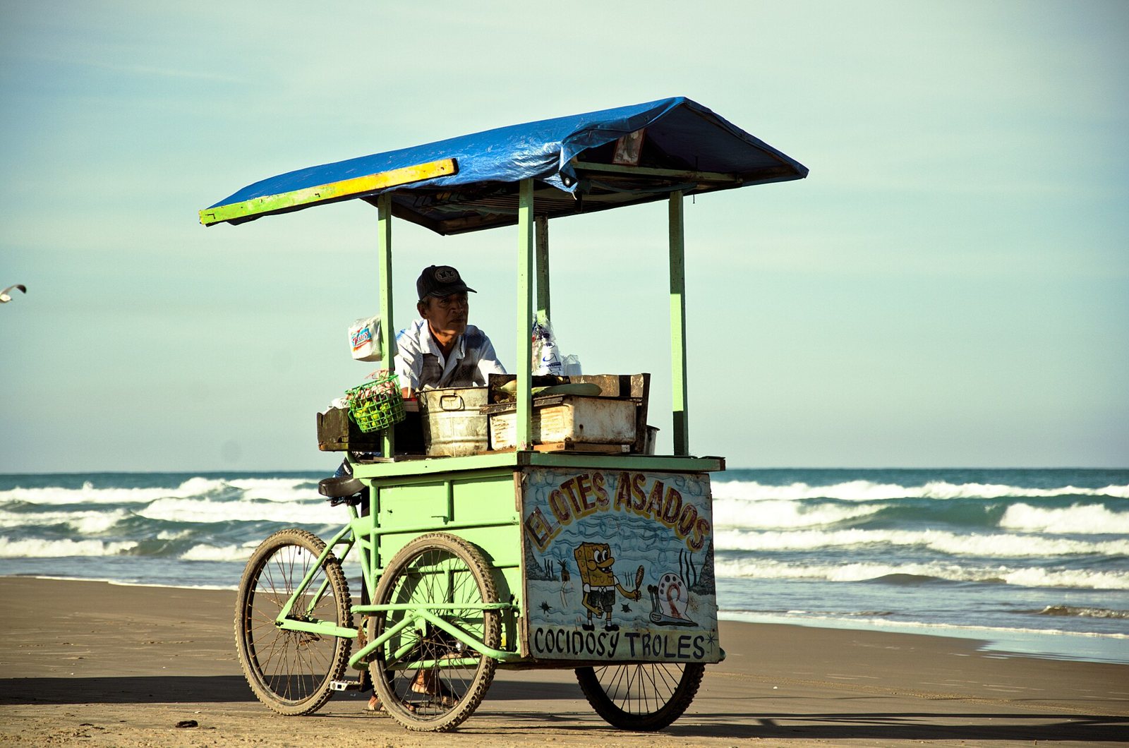 Carrito de elotes asados con toldo azul estacionado sobre la arena, con el mar de fondo