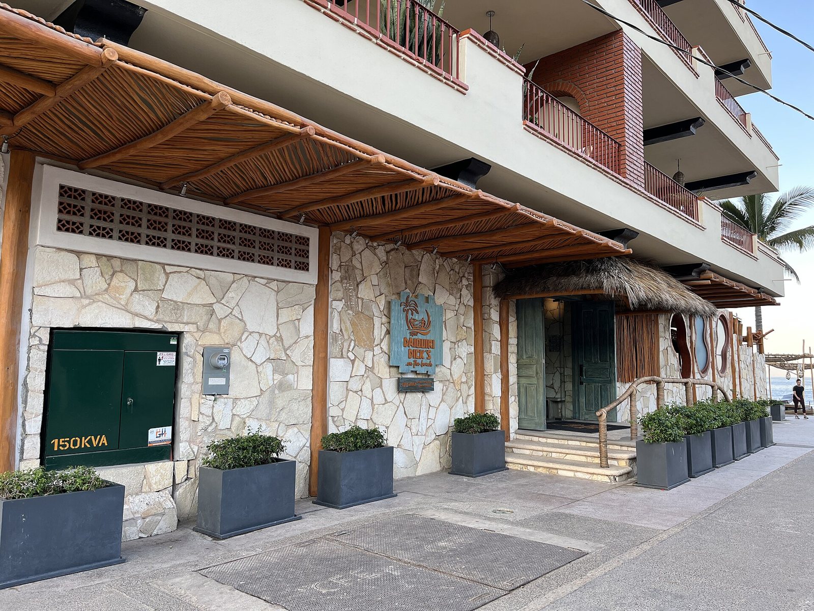 Entrada de Daiquiri Dick's sobre la calle Olas Altas, con muro de piedra, techo de palapa, macetas y el mar al fondo