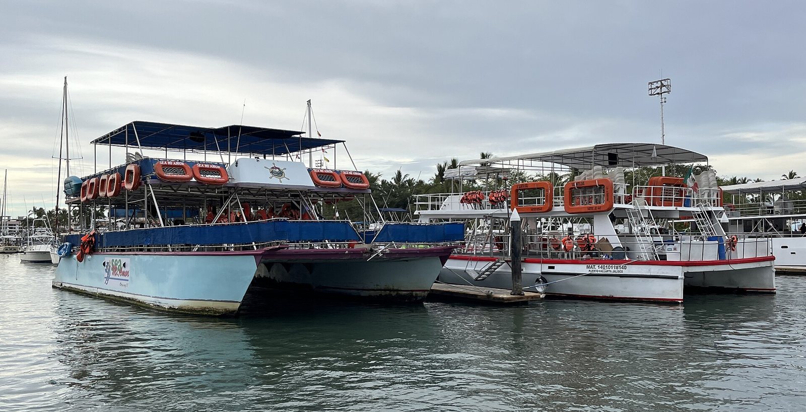 Catamaranes y barcos de paseo con salvavidas naranjas atracados en un muelle de Marina Vallarta en un día nublado