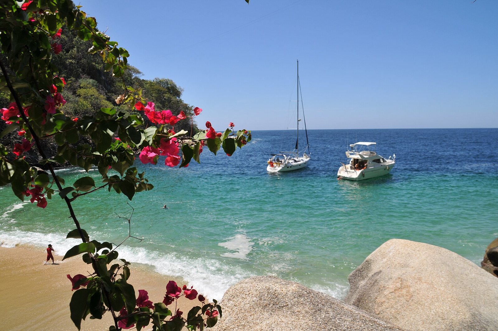 Caleta de arena dorada y agua turquesa con dos embarcaciones ancladas, vista entre flores de bugambilia y rocas de granito